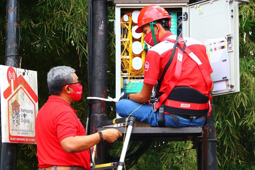  A technician wearing a red uniform and safety gear is repairing a fiber optic cable while another technician is holding a clipboard and looking at the cable.